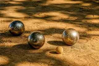 Polished pétanque balls on a sunlit clay court under shade from plane trees, with warm late afternoon light and a tranquil Provençal ambiance.